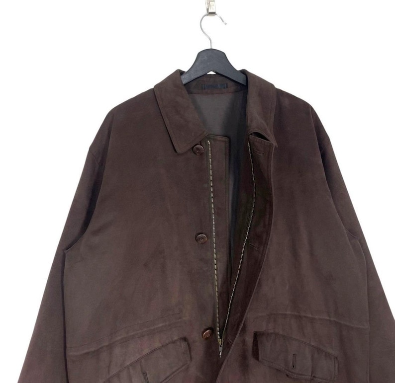 May include: A brown suede jacket with a collared neckline, button closures, and two front pockets. The jacket is hanging on a black hanger against a white background. The jacket appears to be a vintage or pre-owned item.