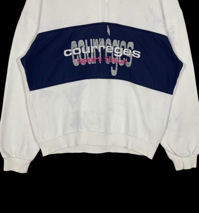 Puede incluir: Sudadera blanca con un panel azul marino en el pecho. El panel presenta el texto bordado "courreges sport futur" en plata y rosa. La sudadera tiene cierre de cremallera de un cuarto y mangas largas.
