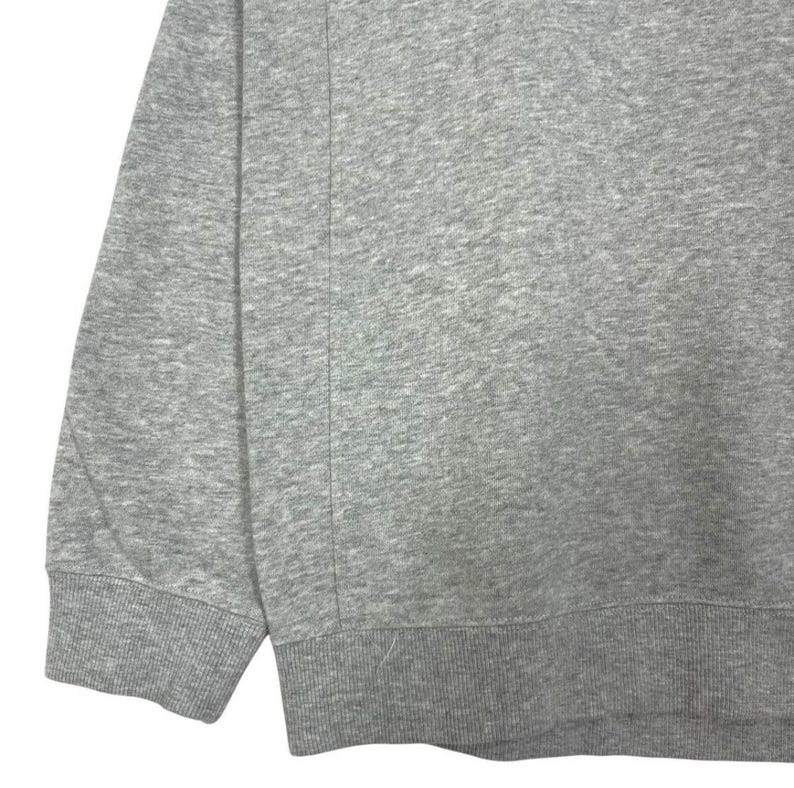 May include: A close-up of a grey sweatshirt. The image shows the sleeve and lower portion of the garment, with a ribbed cuff and hem. The fabric has a textured appearance, and a vertical seam is visible.