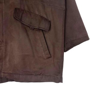 May include: A brown leather jacket with a single pocket on the chest. The jacket has short sleeves and a button closure on the pocket. The leather appears to be worn and has a matte finish.