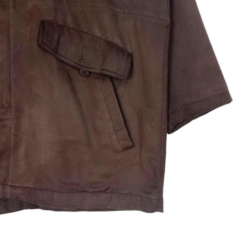 May include: A brown leather jacket with a single pocket on the chest. The jacket has short sleeves and a button closure on the pocket. The leather appears to be worn and has a matte finish.