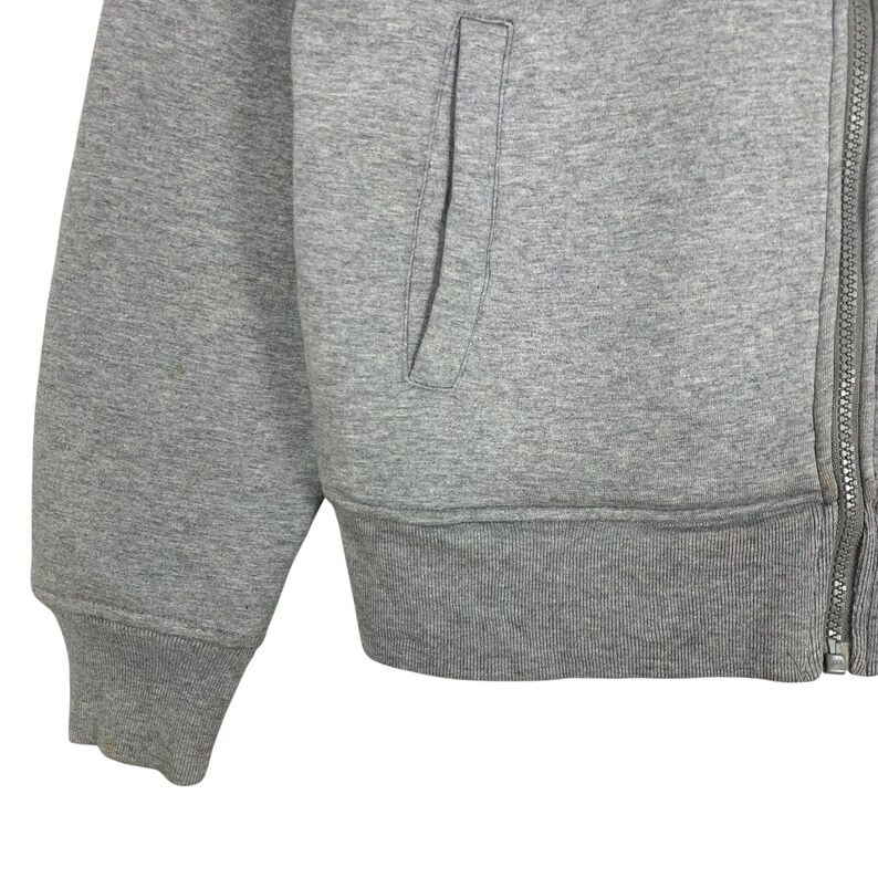 May include: Close-up of a gray zip-up hoodie. The hoodie features a front pocket, ribbed cuffs, and a ribbed hem. The zipper runs down the front of the garment. The fabric appears to be a soft, medium-weight material.