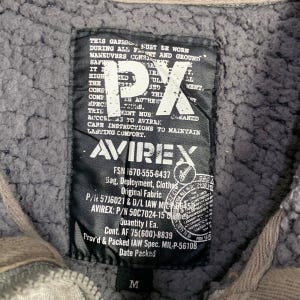 May include: Close-up of a gray jacket label with the text "PX" and "AVIREX". The label includes various text details, including care instructions and specifications. The jacket is gray with a fuzzy interior and a size "M" label.