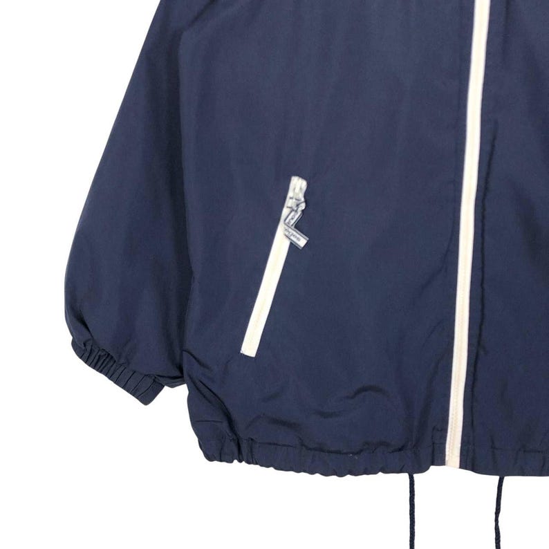 Puede incluir: Chaqueta cortavientos azul marino con cremallera y ribete blancos. La chaqueta tiene un cord&oacute;n en la parte inferior y pu&ntilde;os el&aacute;sticos. Un peque&ntilde;o bolsillo con cremallera blanca es visible en el pecho.