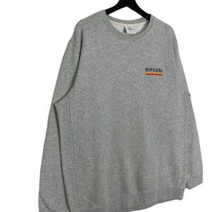 May include: A grey sweatshirt featuring the word "RIPCURL" in orange and blue on the chest. The sweatshirt has a crew neck and long sleeves. The garment is suspended from a black hanger.
