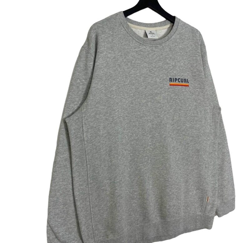 May include: A grey sweatshirt featuring the word "RIPCURL" in orange and blue on the chest. The sweatshirt has a crew neck and long sleeves. The garment is suspended from a black hanger.