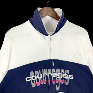 Puede incluir: Sudadera blanca y azul marino con cremallera y el texto "courr&egrave;ges sport futur" en plata y rosa. La sudadera tiene un cuello alto y est&aacute; colgada de una percha de madera.