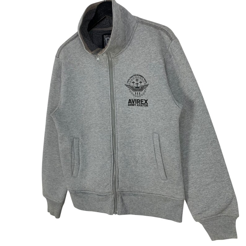 May include: A gray zip-up sweatshirt with a high collar. The sweatshirt features a black graphic on the chest that reads "AVIREX ARMY AVIATOR". The jacket has two front pockets and a full-length zipper.