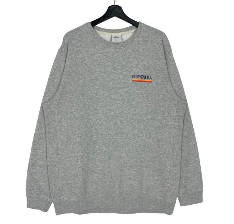 May include: A grey sweatshirt featuring the word "RIPCURL" in orange and white across the chest. The long-sleeved crew neck shirt is made of a soft material.