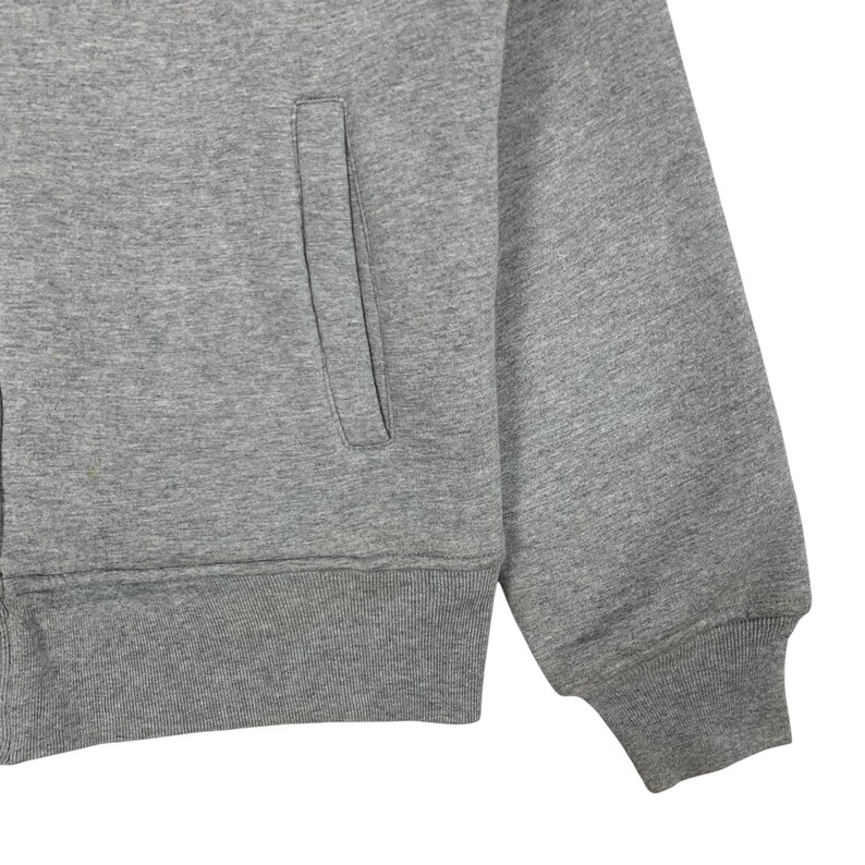 May include: Close-up of a gray zip-up sweatshirt. The sweatshirt features a front pocket and ribbed cuffs and hem. The fabric appears to be a soft, medium-weight material. The sleeve is visible, showing the cuff detail.