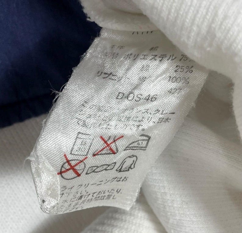 Puede incluir: Primer plano de una etiqueta de tela blanca con texto japon&eacute;s y s&iacute;mbolos de cuidado. La etiqueta incluye el texto "DOS-46" y porcentajes de poli&eacute;ster y algod&oacute;n. La etiqueta est&aacute; adherida a una prenda blanca.