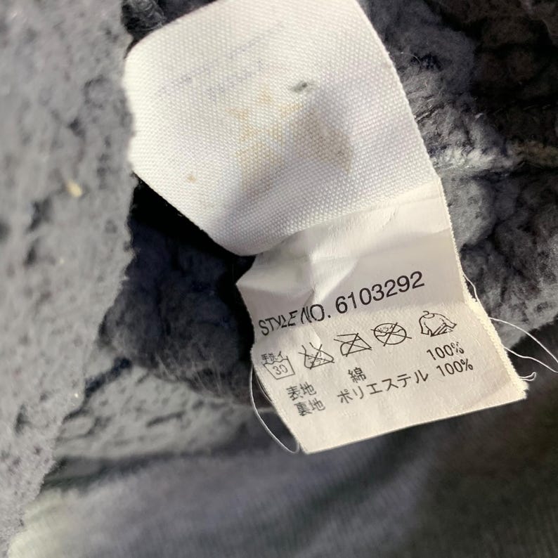 May include: A white fabric tag with the text "STYLE NO. 6103292" and care symbols. The tag is attached to a gray, textured fabric. The tag also includes Japanese text.