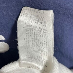 Puede incluir: Primer plano de una etiqueta de tela blanca con texto japon&eacute;s y las palabras "MADE IN JAPAN" impresas en negro. La etiqueta est&aacute; sobre un fondo de tela azul oscuro.