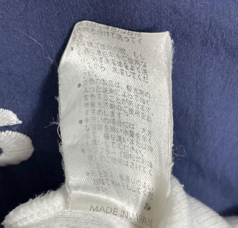 Puede incluir: Primer plano de una etiqueta de tela blanca con texto japon&eacute;s y las palabras "MADE IN JAPAN" impresas en negro. La etiqueta est&aacute; sobre un fondo de tela azul oscuro.