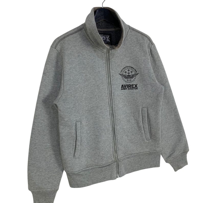 May include: Gray zip-up jacket with a high collar and two front pockets. The jacket features the text "AVIREX ARMY AVIATOR" and a graphic on the upper chest. The jacket has a full zipper closure and a dark gray interior lining.