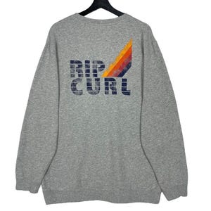 May include: A grey sweatshirt featuring the navy blue text "RIP CURL". A retro design of orange, yellow, red, and blue stripes is on the upper right. The sweatshirt has long sleeves and a crew neck.