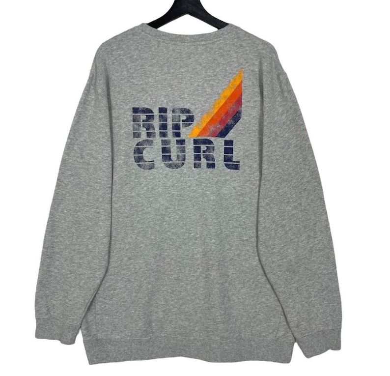 May include: A grey sweatshirt featuring the navy blue text "RIP CURL". A retro design of orange, yellow, red, and blue stripes is on the upper right. The sweatshirt has long sleeves and a crew neck.
