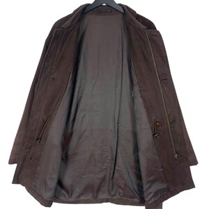 May include: A brown suede coat with a classic design. The coat is open, revealing a dark brown lining. Features include a button closure, a drawstring waist, and a collar. The coat is hanging on a black hanger.