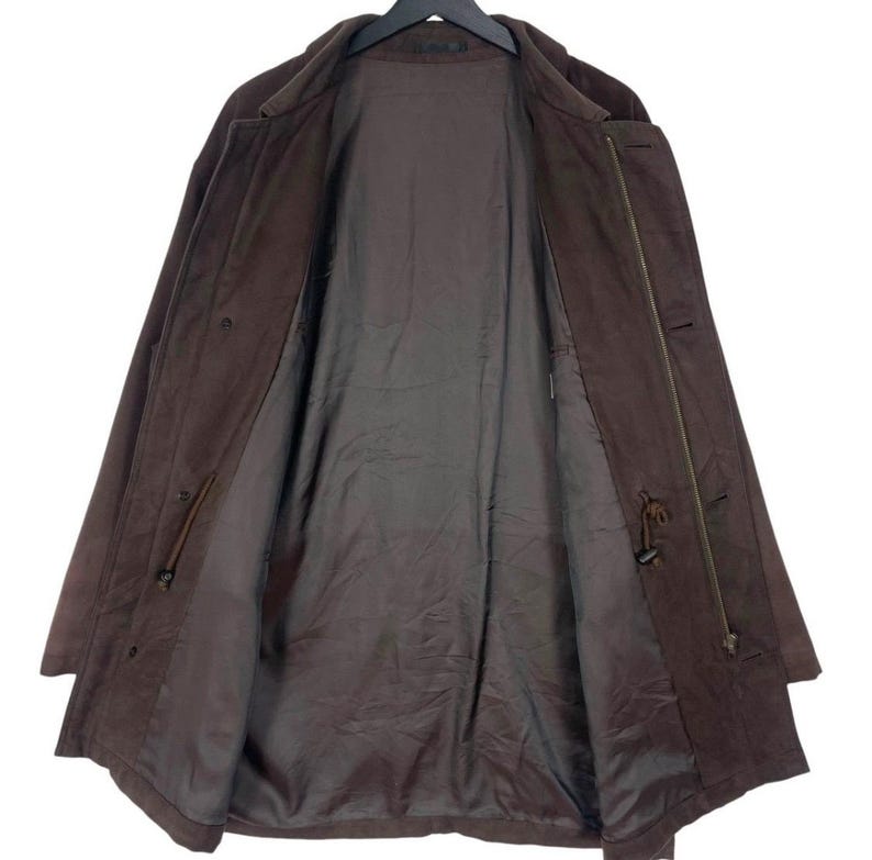 May include: A brown suede coat with a classic design. The coat is open, revealing a dark brown lining. Features include a button closure, a drawstring waist, and a collar. The coat is hanging on a black hanger.