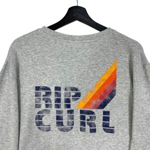 May include: A grey sweatshirt featuring the navy blue text "RIP CURL". A colourful design of orange, red, and yellow stripes is on the upper right. The sweatshirt is suspended from a black hanger.