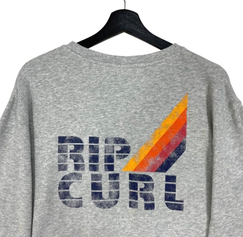 May include: A grey sweatshirt featuring the navy blue text "RIP CURL". A colourful design of orange, red, and yellow stripes is on the upper right. The sweatshirt is suspended from a black hanger.