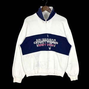 Puede incluir: Sudadera blanca y azul marino con cuello alto y cremallera de cuarto. La sudadera presenta una franja horizontal azul marino en el pecho con el texto "courreges sport club" en fuente plateada. La parte superior de manga larga cuelga sobre un fondo negro.