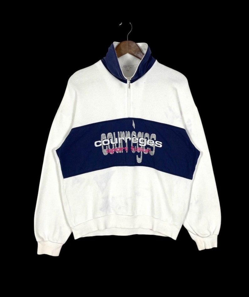 Puede incluir: Sudadera blanca y azul marino con cuello alto y cremallera de cuarto. La sudadera presenta una franja horizontal azul marino en el pecho con el texto "courreges sport club" en fuente plateada. La parte superior de manga larga cuelga sobre un fondo negro.