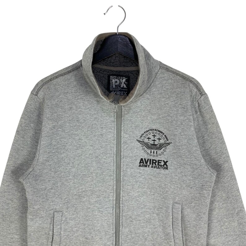 May include: A gray zip-up sweatshirt with a high collar. The sweatshirt features a black graphic on the chest that reads "15th Fighter Bomber Wing" and "Avirex Army Aviator." The brand logo "PX Avirex" is on the collar.