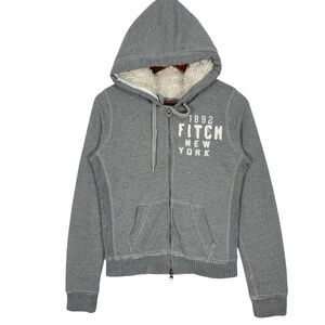 Könnte beinhalten: Ein grauer Kapuzenpullover mit Reißverschluss und einer mit cremefarbenem Kunstfell gefütterten Kapuze. Der Hoodie trägt den weißen Schriftzug "1892 FITCH NEW YORK" auf der Brust. Er hat einen Kordelzug und zwei Vordertaschen.