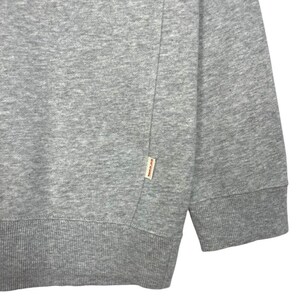 May include: Close-up of a grey sweatshirt. The sweatshirt features a ribbed hem and cuffs. A small rectangular tag is sewn on the lower left side. The fabric appears to be a soft, medium-weight material.