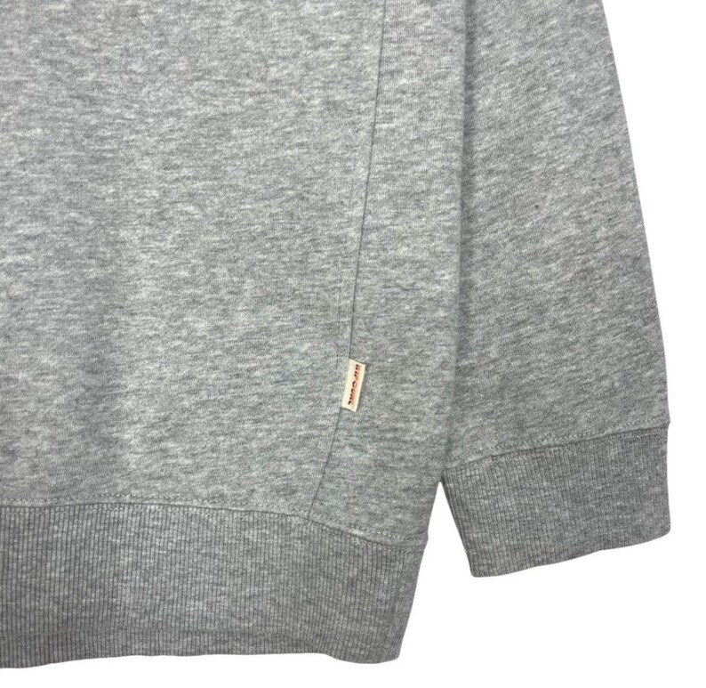 May include: Close-up of a grey sweatshirt. The sweatshirt features a ribbed hem and cuffs. A small rectangular tag is sewn on the lower left side. The fabric appears to be a soft, medium-weight material.