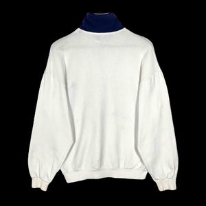 Puede incluir: Una sudadera blanca con cuello alto azul marino. La parte superior de manga larga tiene un corte holgado y pu&ntilde;os acanalados. La vista trasera de la prenda se muestra sobre un fondo negro.