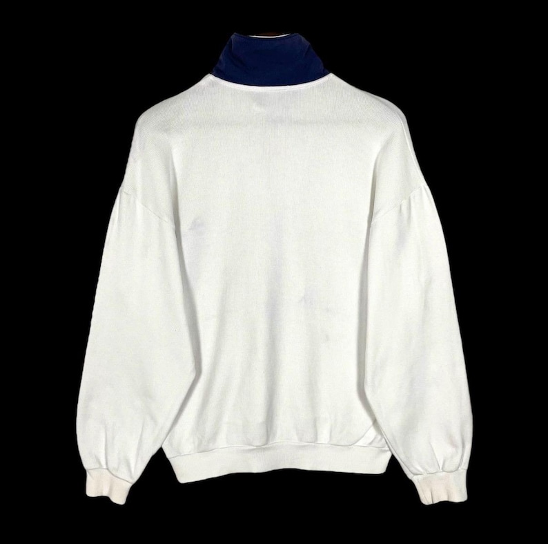 Puede incluir: Una sudadera blanca con cuello alto azul marino. La parte superior de manga larga tiene un corte holgado y pu&ntilde;os acanalados. La vista trasera de la prenda se muestra sobre un fondo negro.