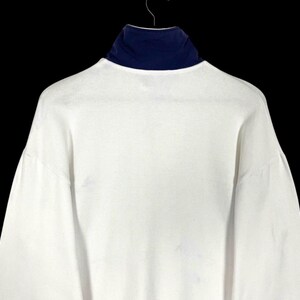 Puede incluir: Una camisa blanca de manga larga con cuello alto azul marino. La camisa est&aacute; colgada de una percha de metal sobre un fondo negro. La camisa tiene una textura acanalada.