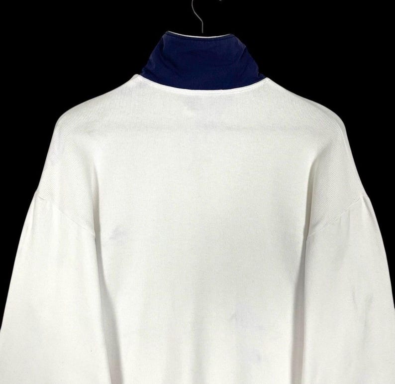 Puede incluir: Una camisa blanca de manga larga con cuello alto azul marino. La camisa est&aacute; colgada de una percha de metal sobre un fondo negro. La camisa tiene una textura acanalada.