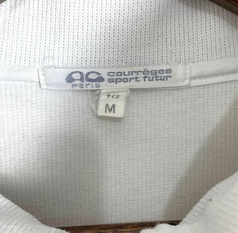 Puede incluir: Primer plano de la etiqueta de una camisa blanca con cuello. La etiqueta presenta la marca "courr&egrave;ges sport futur" y la talla "M". La etiqueta tambi&eacute;n incluye el texto "Paris" y caracteres japoneses.