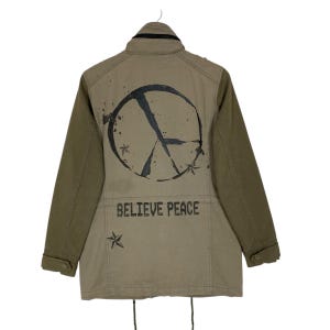 May include: An olive green jacket with long sleeves, featuring a black peace symbol graphic on the back. The words "BELIEVE PEACE" are printed below. The jacket has a button-up front and a drawstring at the bottom.