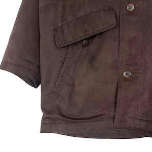 May include: A brown short-sleeved jacket with two front pockets and button closures. The jacket has a classic design and appears to be made of a smooth, possibly leather-like material. The buttons are a matching brown color.