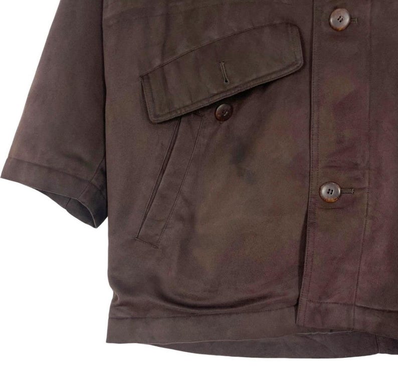 May include: A brown short-sleeved jacket with two front pockets and button closures. The jacket has a classic design and appears to be made of a smooth, possibly leather-like material. The buttons are a matching brown color.