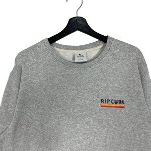 May include: A grey sweatshirt with a crew neck. The word "RIPCURL" is embroidered in a block font across the chest, with a horizontal stripe design in orange, red, and yellow. The sweatshirt is hanging on a black hanger.