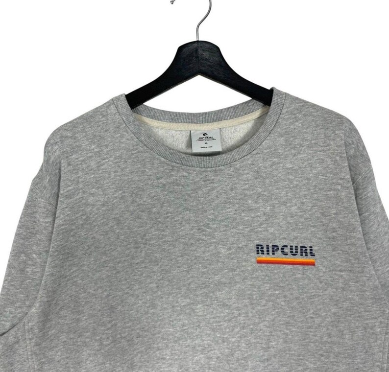 May include: A grey sweatshirt with a crew neck. The word "RIPCURL" is embroidered in a block font across the chest, with a horizontal stripe design in orange, red, and yellow. The sweatshirt is hanging on a black hanger.