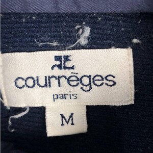 Puede incluir: Primer plano de una etiqueta blanca con el nombre de la marca "courr&egrave;ges paris" en azul. Debajo, la letra "M" est&aacute; escrita en un rect&aacute;ngulo blanco. La etiqueta est&aacute; adherida a una tela azul marino.