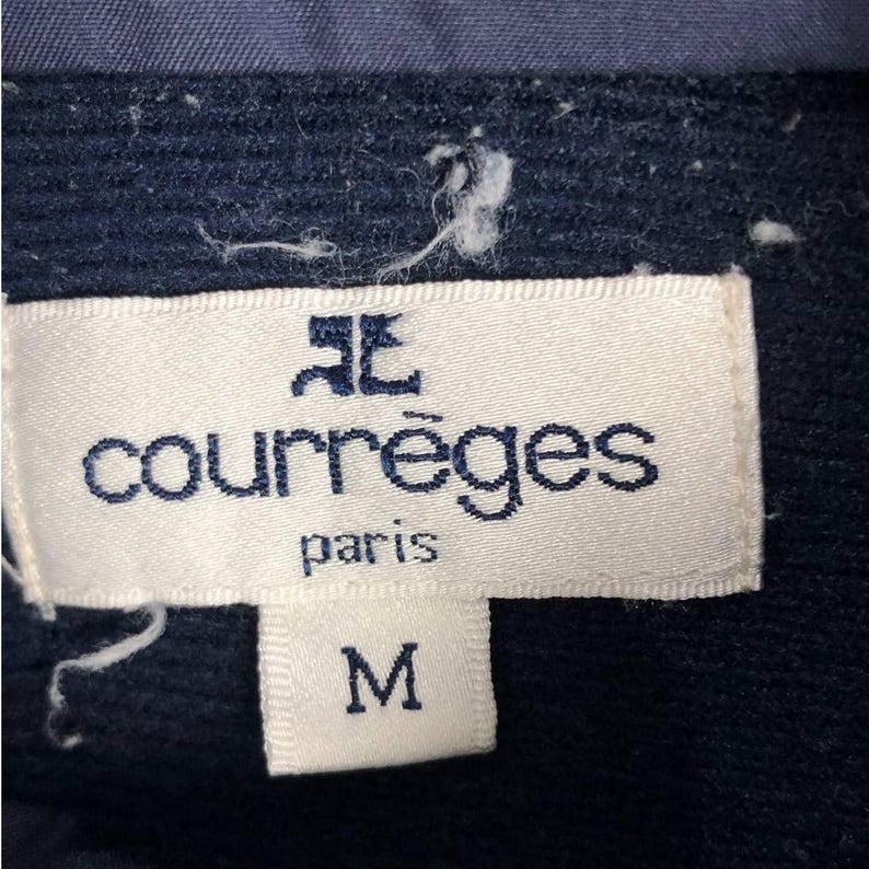 Puede incluir: Primer plano de una etiqueta blanca con el nombre de la marca "courr&egrave;ges paris" en azul. Debajo, la letra "M" est&aacute; escrita en un rect&aacute;ngulo blanco. La etiqueta est&aacute; adherida a una tela azul marino.
