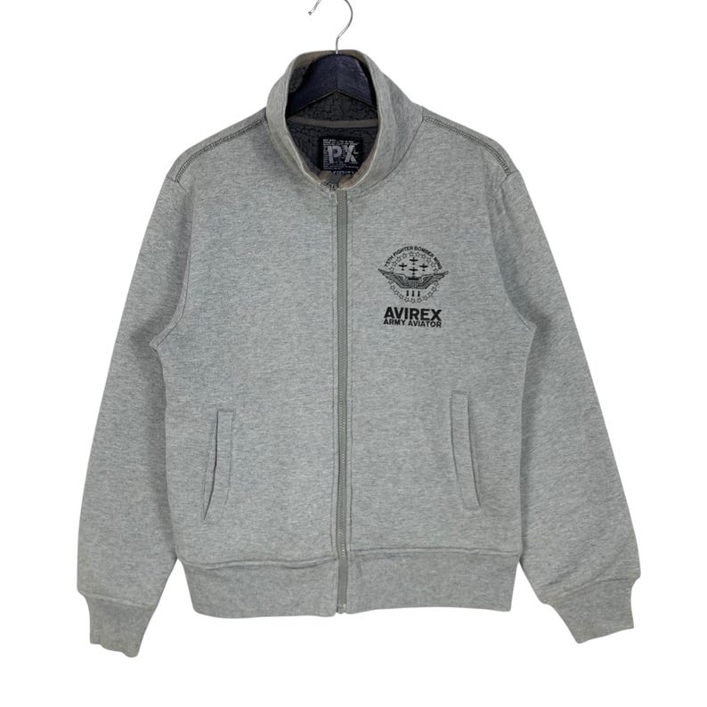 May include: A gray zip-up sweatshirt with a stand-up collar. The sweatshirt features the text "PX" on the collar and a graphic with the text "AVIREX ARMY AVIATOR" on the chest. It has two front pockets.