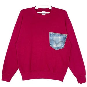 Könnte beinhalten: Ein weinrotes Sweatshirt mit einer Jeans-Tasche. Die Tasche ist aus hellblauem Denim mit verwaschenem Aussehen. Das Sweatshirt hängt an einem weißen Kleiderbügel.