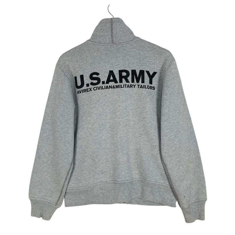 May include: A gray zip-up sweatshirt with the text "U.S.ARMY" in large black letters across the back. Below the text is the text "AVIREX CIVILIAN&MILITARY TAILORS" in smaller black letters. The sweatshirt has a high collar.