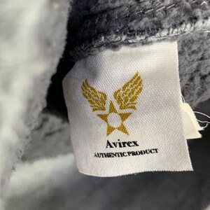 May include: Close-up of a white label with the Avirex logo, featuring a star and wings design in gold. The text "Avirex" and "AUTHENTIC PRODUCT" are printed in black. The label is attached to a gray fabric.