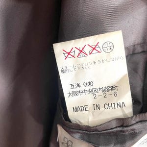 May include: Close-up of a clothing tag with Japanese text and care symbols. The tag reads "MADE IN CHINA" in English. The background is a dark gray fabric.