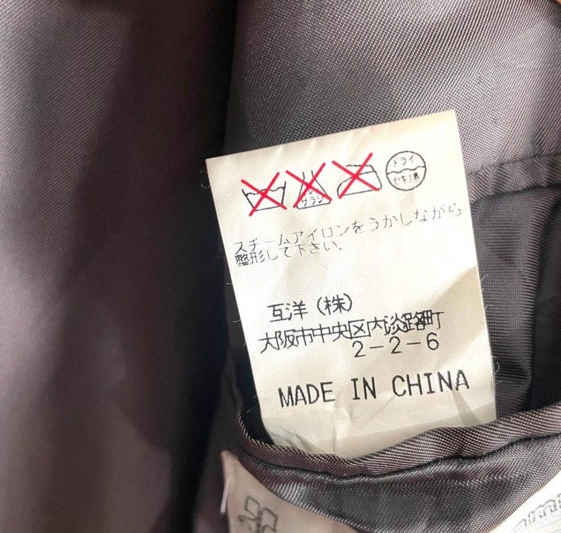 May include: Close-up of a clothing tag with Japanese text and care symbols. The tag reads "MADE IN CHINA" in English. The background is a dark gray fabric.