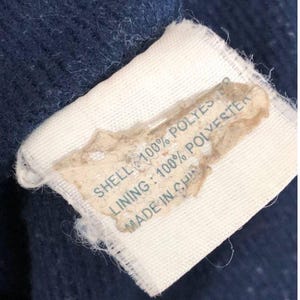 Puede incluir: Primer plano de una etiqueta sobre una tela azul marino. La etiqueta es blanca con una secci&oacute;n beige. El texto de la etiqueta dice "SHELL: 100% POLYESTER", "LINING: 100% POLYESTER" y "MADE IN CHINA".